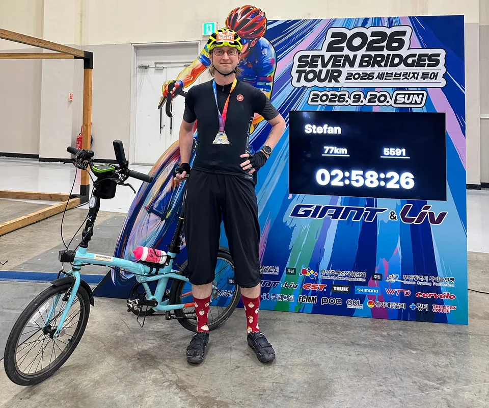 Stefan wearing a finisher medal beside his folding bike and the Seven Bridges 2026 finisher display